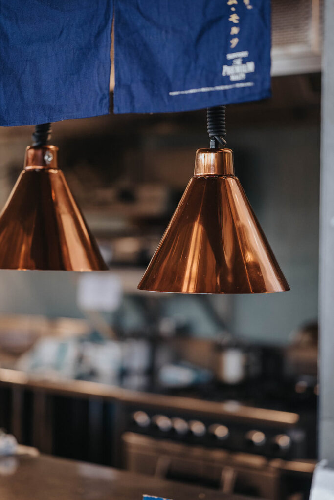 Ample stainless steel work benches, Brass lighting and upholstered seating and complementary wooden chairs for this hospitality fit out for Nippon Izakaya Bar and Kitchen, Total Fitouts Perth South